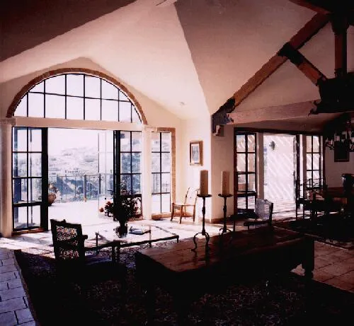 Spacious living room with large arched windows, wooden beams, and elegant furnishings. Sunlight illuminates the space, casting shadows on the tiled floor. Spacious living room with large arched windows, wooden beams, and elegant furnishings. Sunlight illuminates the space, casting shadows on the tiled floor.