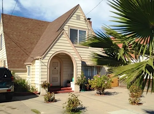 A small house with a steep gable roof and an arched entrance, surrounded by a driveway, shrubs, and a palm tree. A small house with a steep gable roof and an arched entrance, surrounded by a driveway, shrubs, and a palm tree.