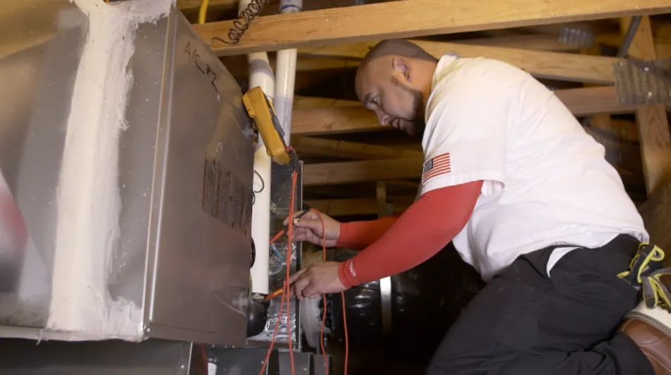 Technician kneeling in attic, checking wiring on a furnace using tools. Wooden beams and ductwork visible overhead. Technician kneeling in attic, checking wiring on a furnace using tools. Wooden beams and ductwork visible overhead.