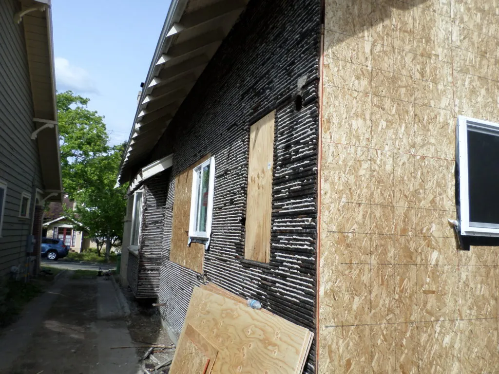 Partial view of a house under renovation, with new plywood siding and exposed black building paper. Tools and materials are visible in the foreground. Partial view of a house under renovation, with new plywood siding and exposed black building paper. Tools and materials are visible in the foreground.