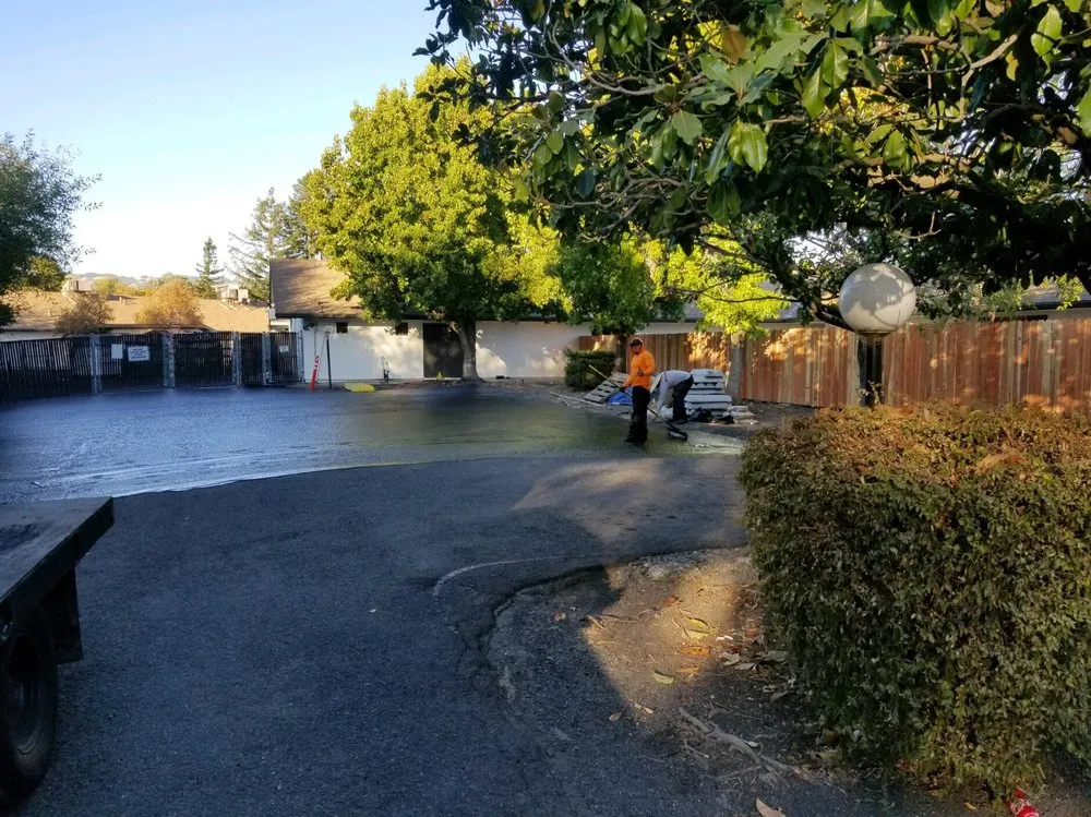 Workers spread asphalt in a shaded outdoor area, surrounded by trees and a wooden fence. Workers spread asphalt in a shaded outdoor area, surrounded by trees and a wooden fence.