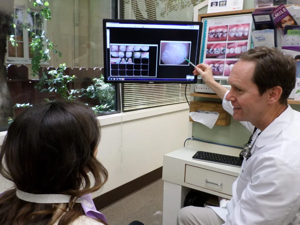 Dentist pointing to dental scans on a monitor while explaining to a patient. The room has a window with greenery outside. Dentist pointing to dental scans on a monitor while explaining to a patient. The room has a window with greenery outside.