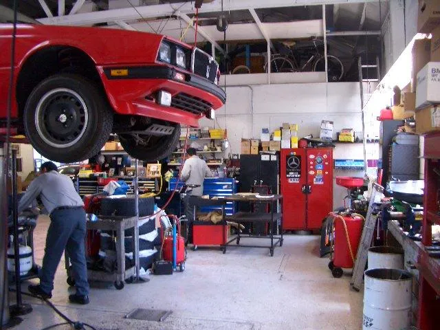 A red car is elevated on a lift in a busy auto repair shop with technicians working and tools organized around the space. A red car is elevated on a lift in a busy auto repair shop with technicians working and tools organized around the space.
