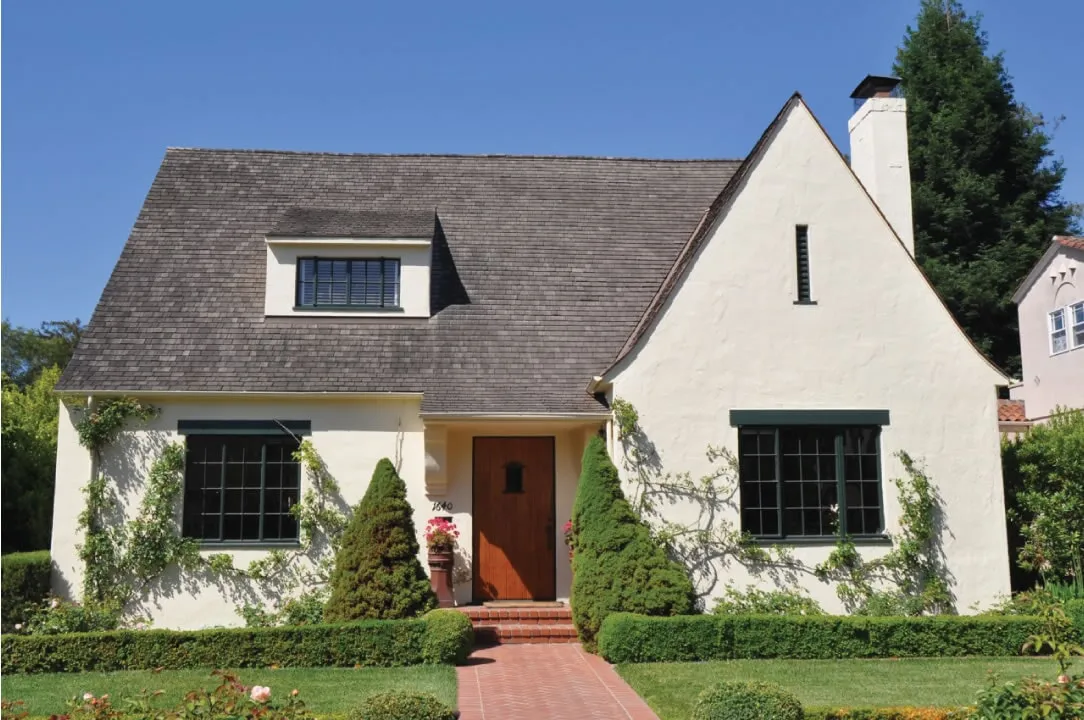 Front view of a single-story house with a steep roof, large windows, white walls, and surrounded by well-maintained greenery and shrubs. Front view of a single-story house with a steep roof, large windows, white walls, and surrounded by well-maintained greenery and shrubs.
