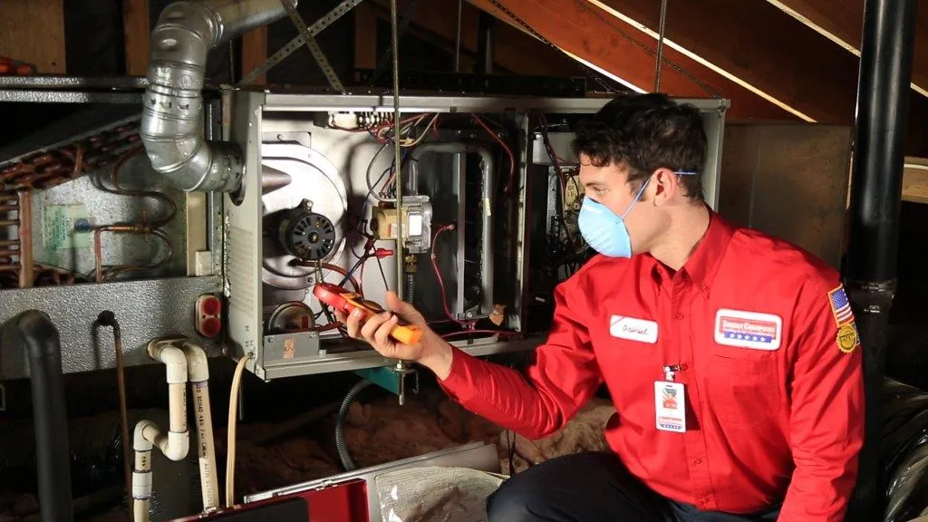Technician in red uniform uses a multimeter to check a furnace in an attic, wearing a face mask. Tools and wiring are visible. Technician in red uniform uses a multimeter to check a furnace in an attic, wearing a face mask. Tools and wiring are visible.