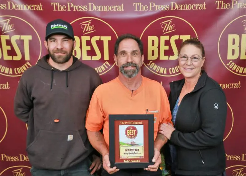  Three people posing in front of a backdrop, holding an award plaque for "Best Electrician."