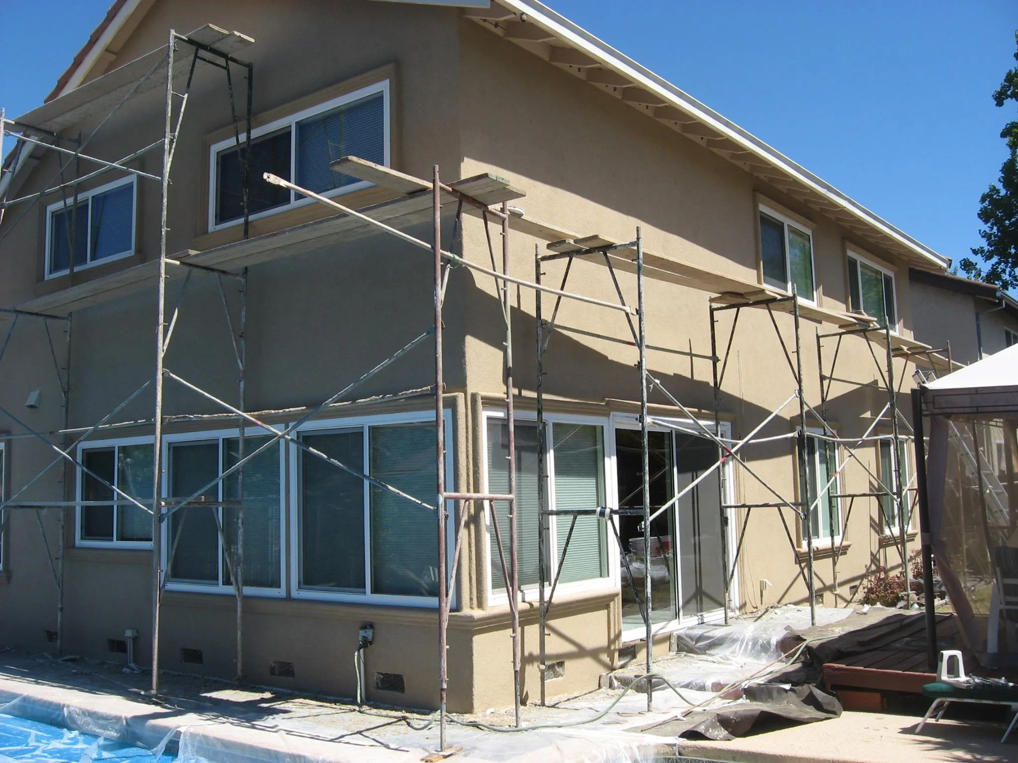 Scaffolding surrounds a beige two-story house. Windows are visible, with clear skies above. Plastic sheets cover the ground, indicating ongoing exterior work. Scaffolding surrounds a beige two-story house. Windows are visible, with clear skies above. Plastic sheets cover the ground, indicating ongoing exterior work.