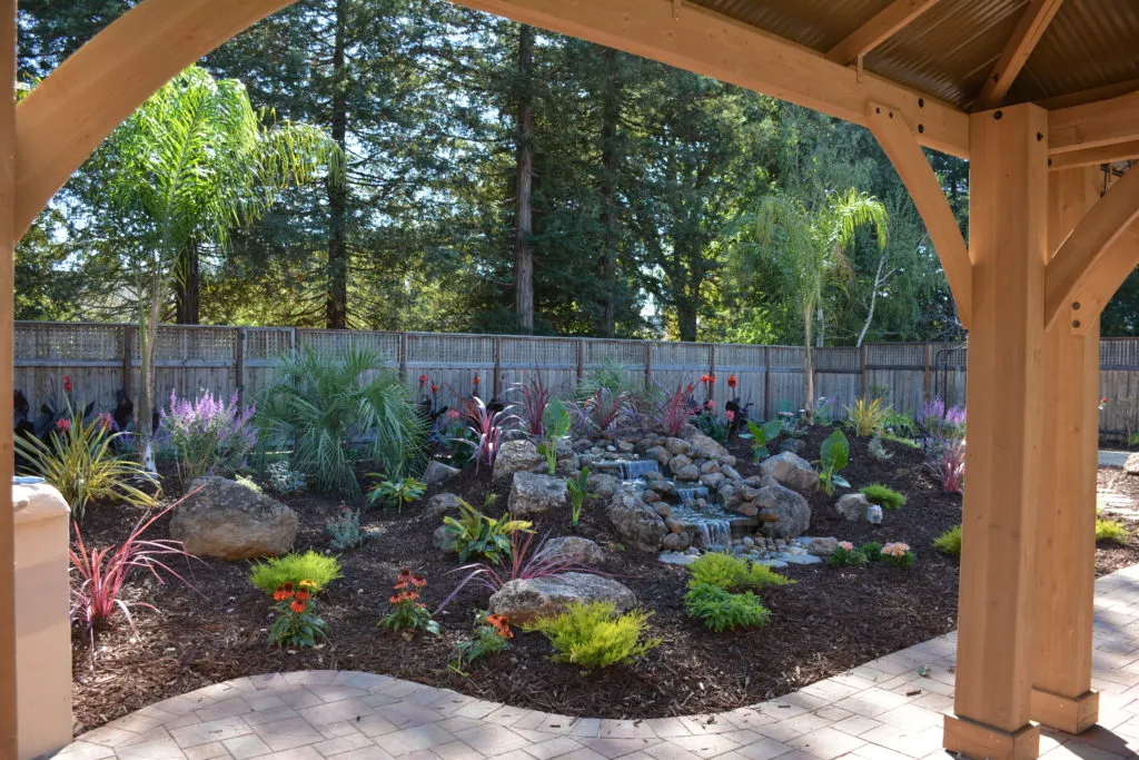  Garden with a water feature, rocks, colorful plants, and trees, viewed from under a wooden pergola.