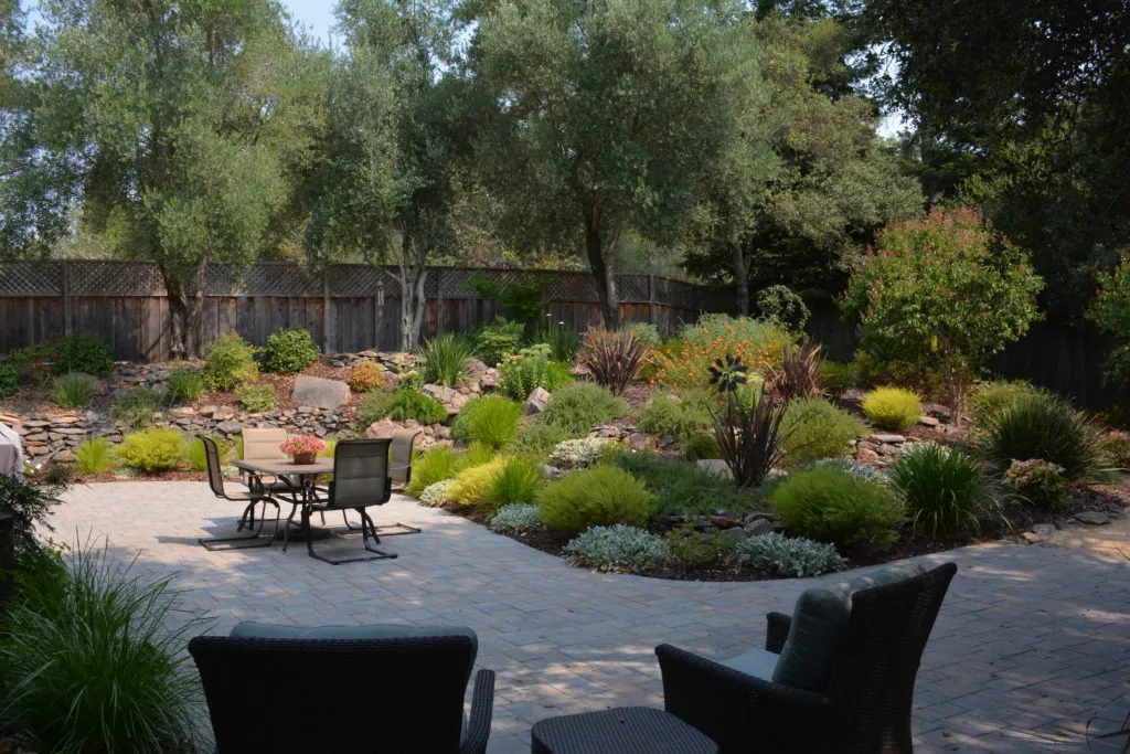  A landscaped garden with a stone patio, surrounded by various shrubs and trees, and outdoor furniture in the foreground.