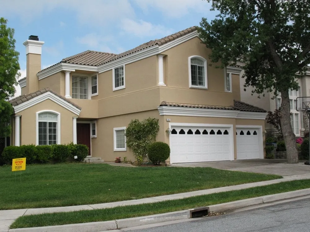  Two-story beige house with white trim and a three-car garage, surrounded by green lawn and shrubs, under a clear sky. A sign reading "Faith Painting" is visible on the grass.