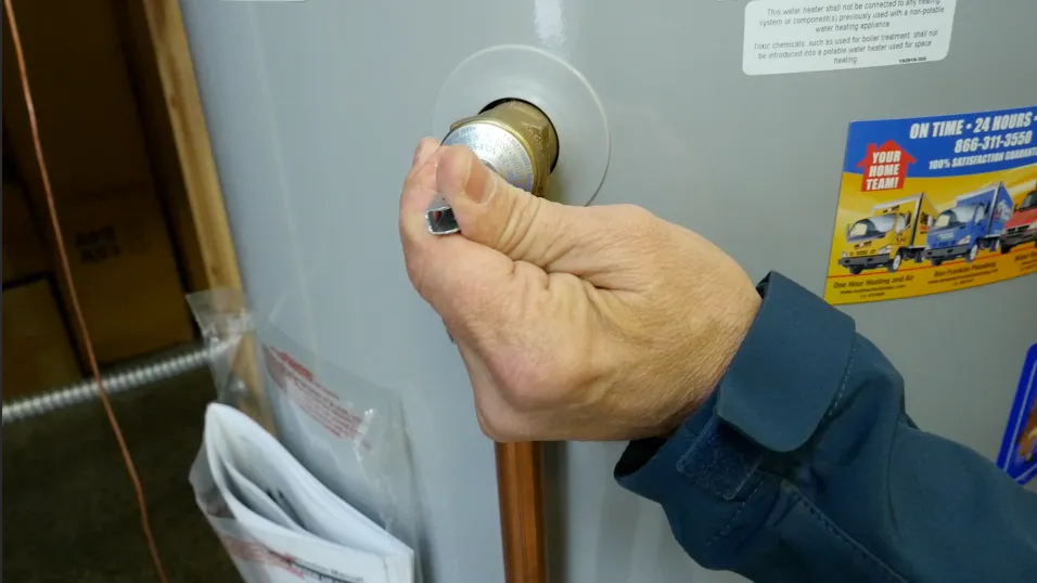 A hand in a blue sleeve adjusts a fitting on a water heater, with copper piping visible and informational stickers attached to the unit. A hand in a blue sleeve adjusts a fitting on a water heater, with copper piping visible and informational stickers attached to the unit.