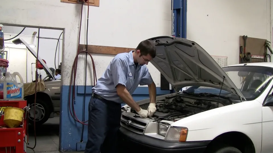 Technician working under the hood of a white car in a garage, surrounded by tools and automotive supplies. Technician working under the hood of a white car in a garage, surrounded by tools and automotive supplies.