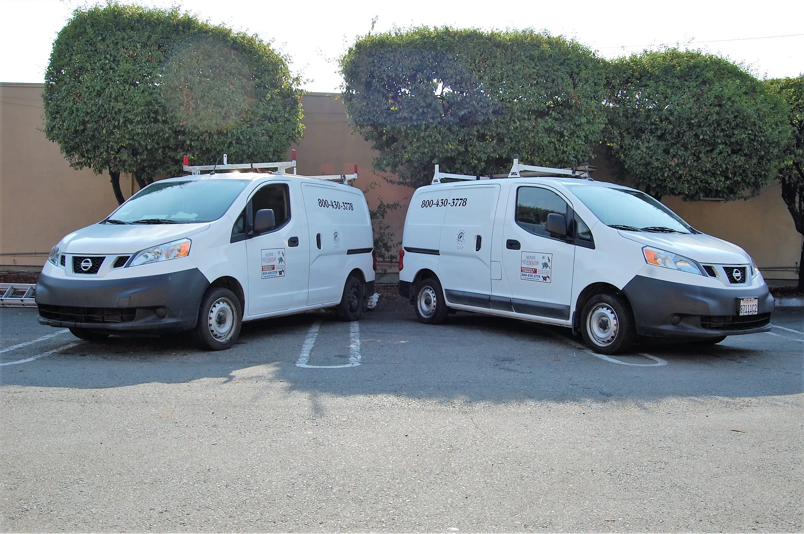  Two white vans with company logos and contact numbers parked side by side in a parking lot in front of trimmed bushes.
