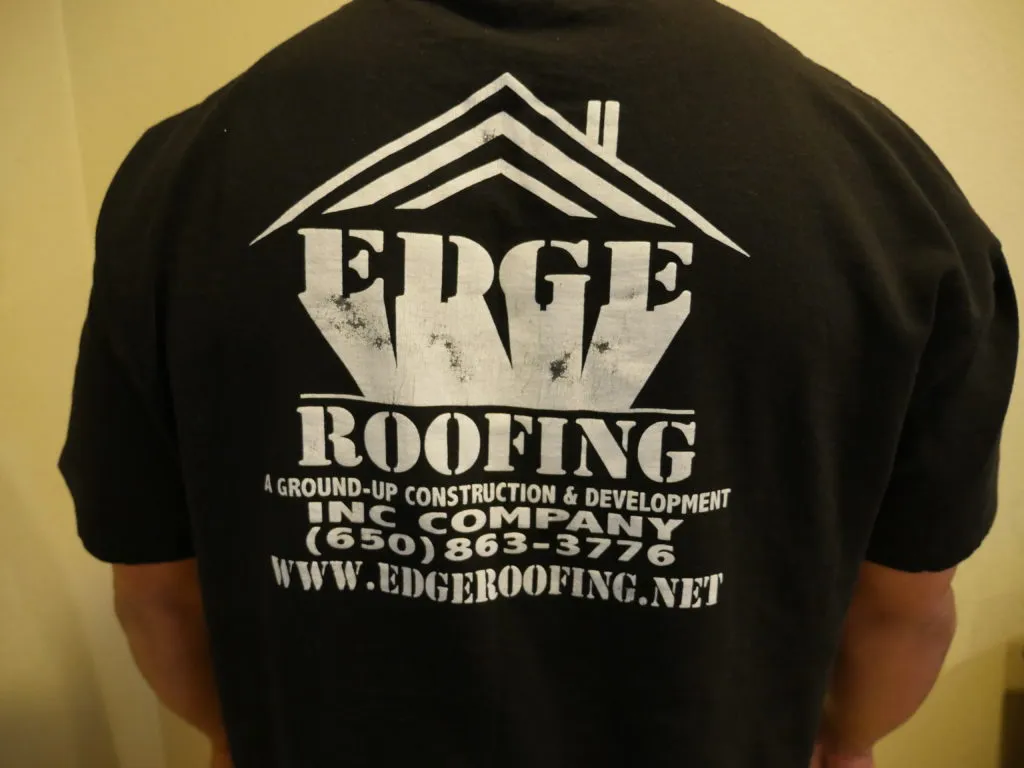 Close-up of a person's back wearing a black shirt with the Edge Roofing logo and contact information printed on it. Close-up of a person's back wearing a black shirt with the Edge Roofing logo and contact information printed on it.
