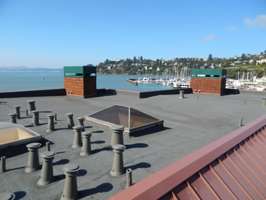 Flat roof with multiple vents, a skylight, and two boxy structures, overlooking a marina with boats and distant hills. Flat roof with multiple vents, a skylight, and two boxy structures, overlooking a marina with boats and distant hills.