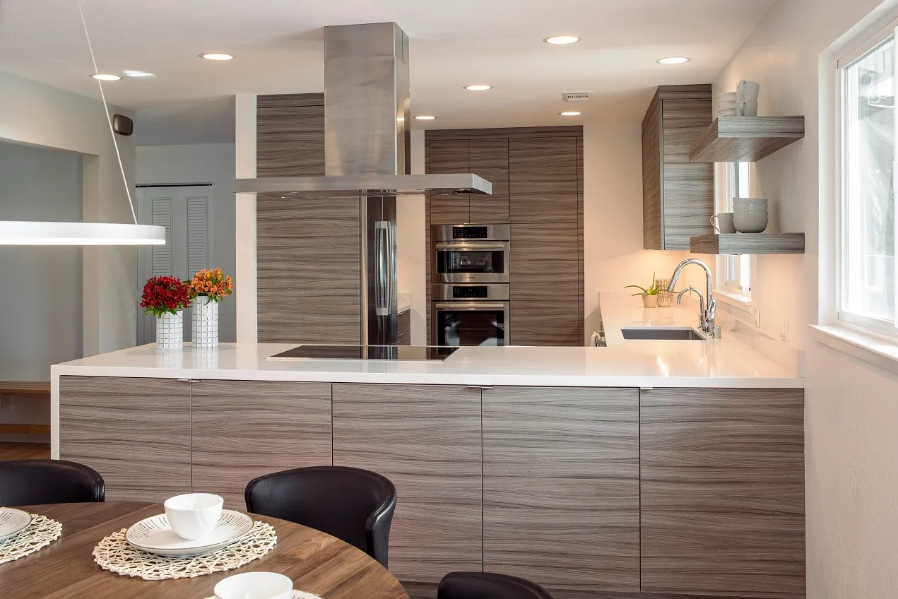 Modern kitchen with sleek wooden cabinets, central island with stovetop, overhead hood, double oven, and open shelving with decor.