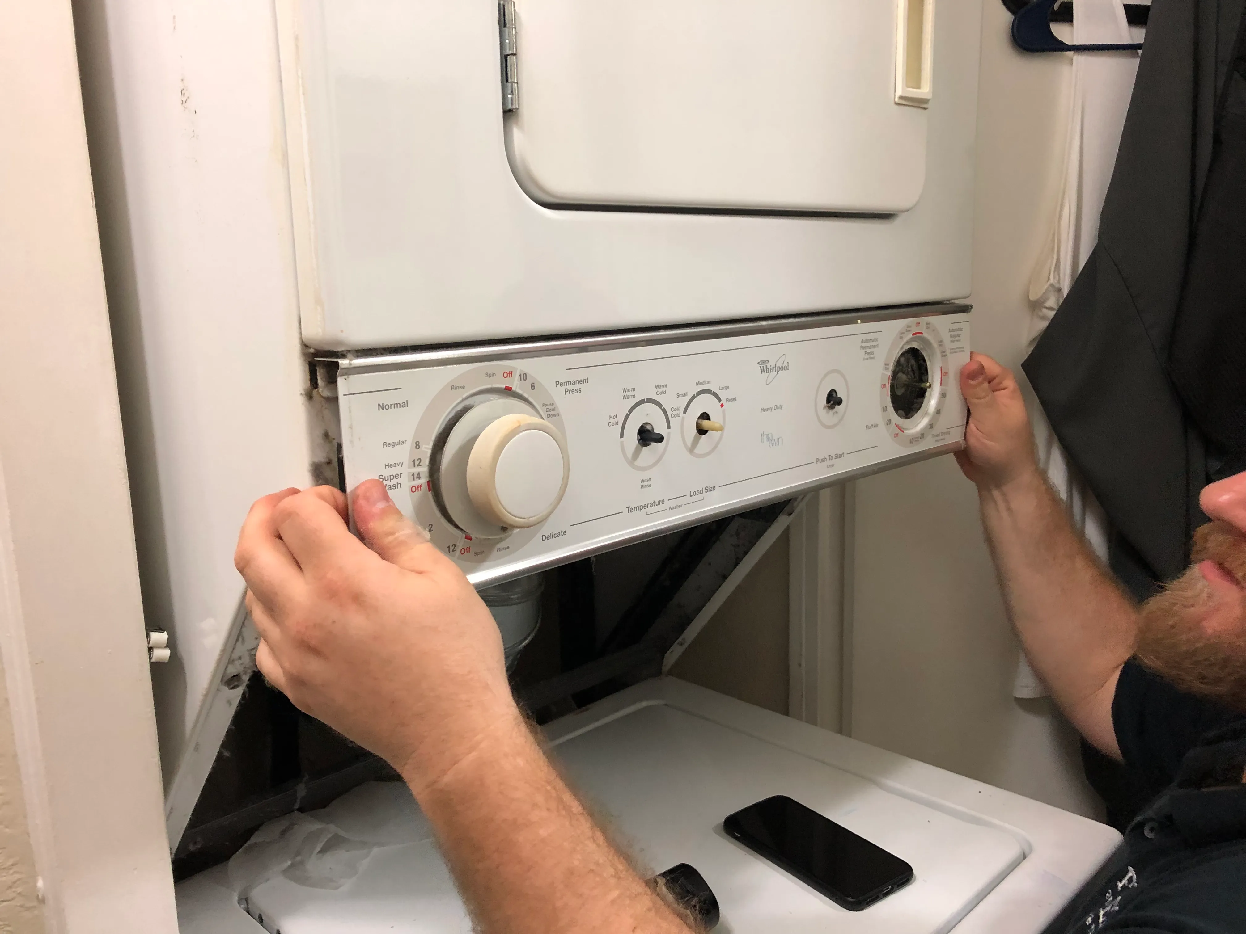 A person is adjusting the control panel of a stacked laundry unit. A smartphone rests on the lower machine. A person is adjusting the control panel of a stacked laundry unit. A smartphone rests on the lower machine.