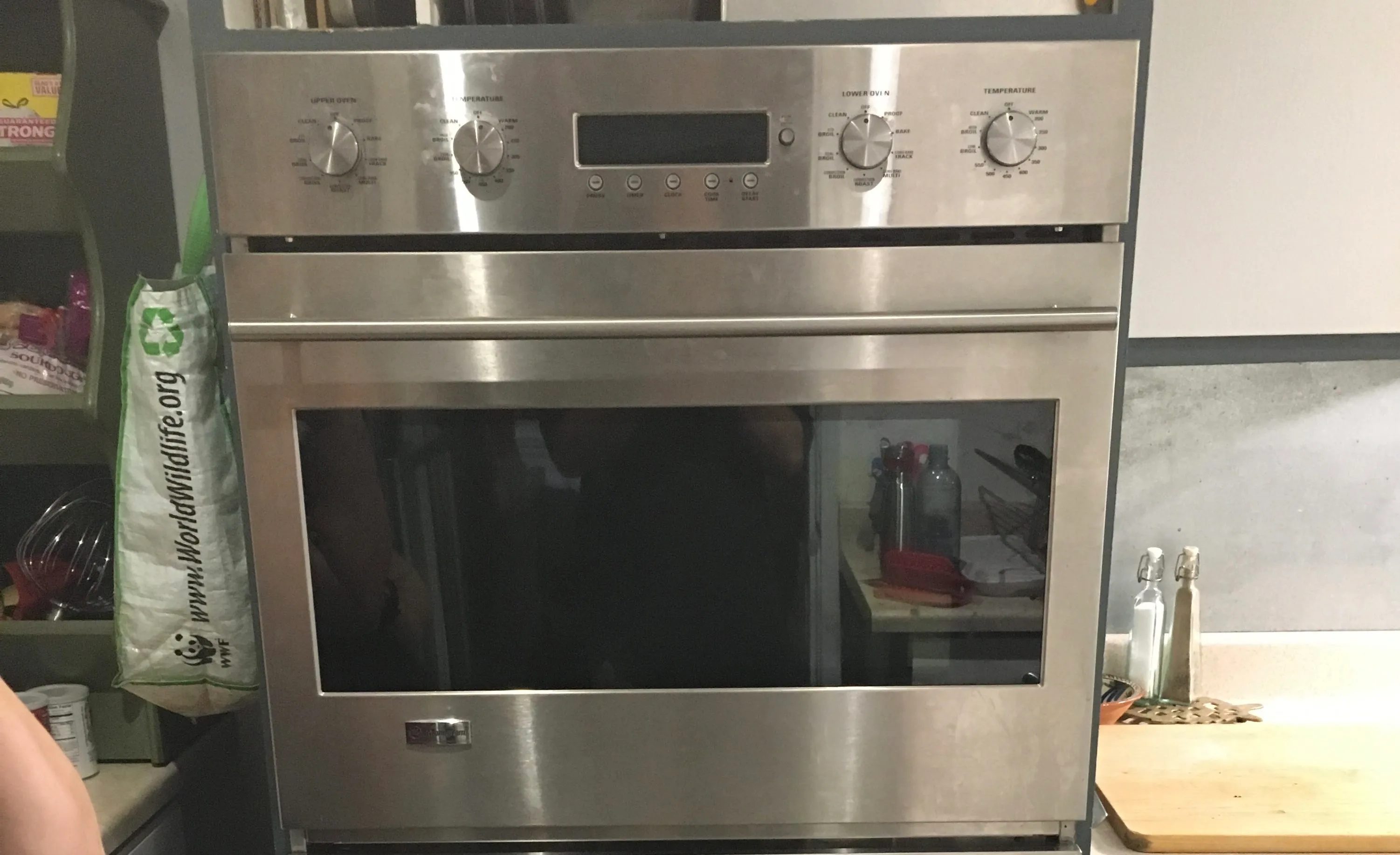 Stainless steel double oven with control knobs displayed, surrounded by kitchen items and a recycling bag on the side.