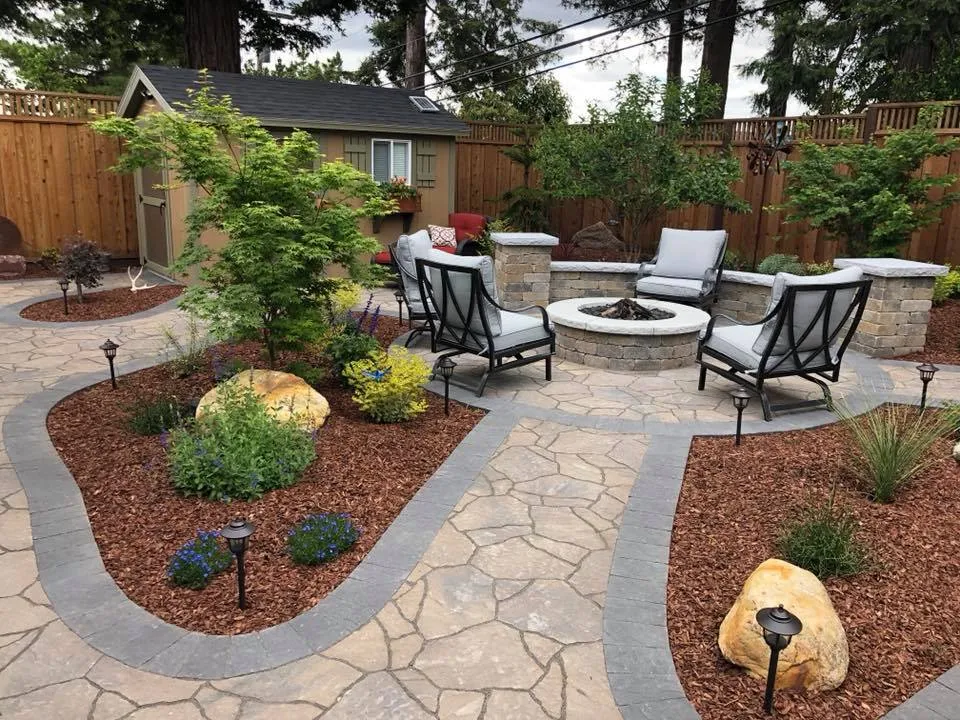 Stone patio with a fire pit surrounded by chairs, bordered by landscaped garden beds and mulch. Stone patio with a fire pit surrounded by chairs, bordered by landscaped garden beds and mulch.