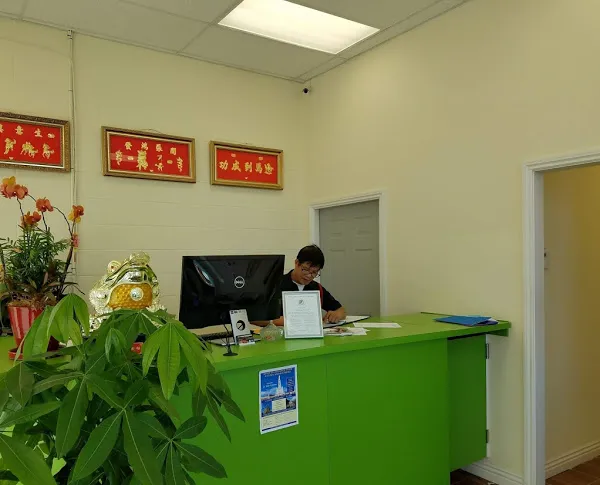   
A staff member sits at a green reception desk, working with papers. A computer and a plant are on the desk. Wall decorations and a door are in the background.