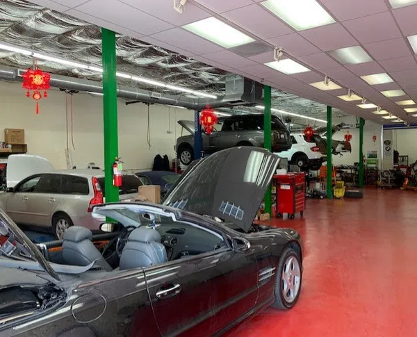 Interior of an auto repair shop with cars on lifts, including a convertible with an open hood, red toolboxes nearby, and visible overhead ductwork.