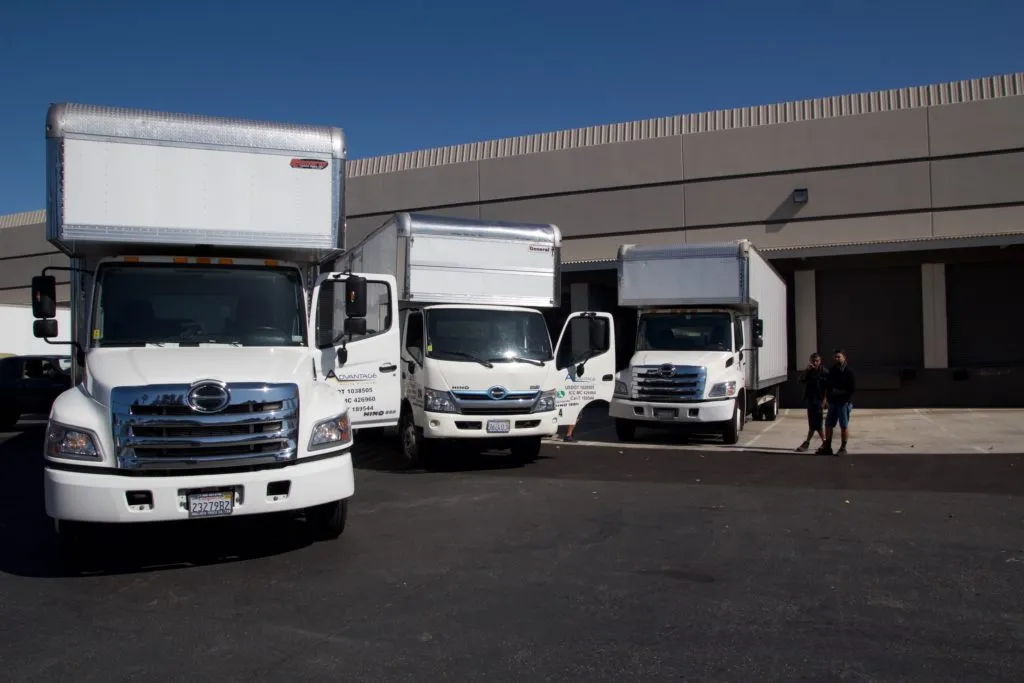 Three white moving trucks parked in front of a warehouse, with two people standing nearby on a sunny day. Three white moving trucks parked in front of a warehouse, with two people standing nearby on a sunny day.