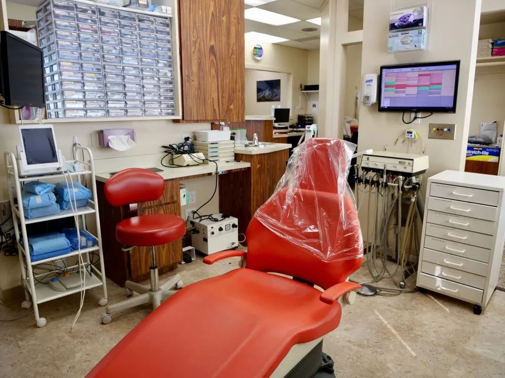 Dental office with a red chair, dental tools, monitor displaying a schedule, storage drawers, and an organized supply area. Dental office with a red chair, dental tools, monitor displaying a schedule, storage drawers, and an organized supply area.