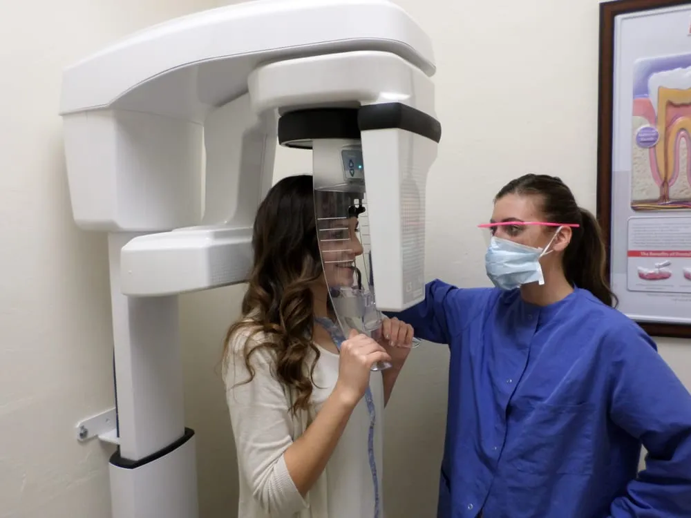 A patient is positioned in a dental scanning machine while a technician wearing a mask and protective glasses observes. A patient is positioned in a dental scanning machine while a technician wearing a mask and protective glasses observes.