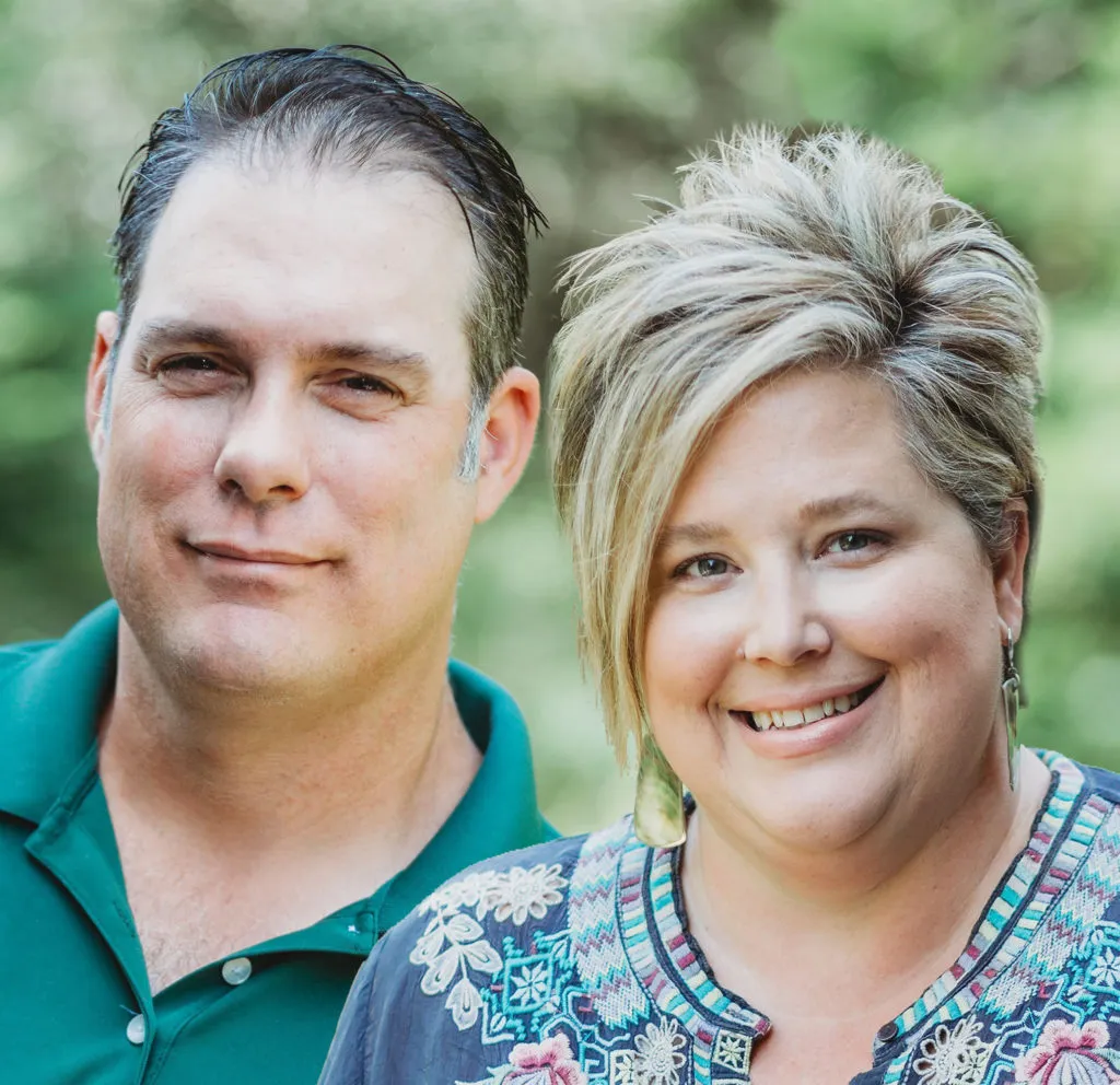 Two people, a man and a woman, smiling outdoors with greenery in the background.