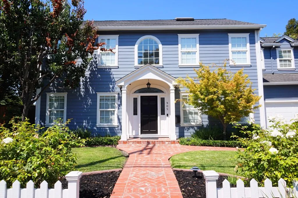 Blue two-story house with white trim, surrounded by green foliage and a white picket fence. Blue two-story house with white trim, surrounded by green foliage and a white picket fence.