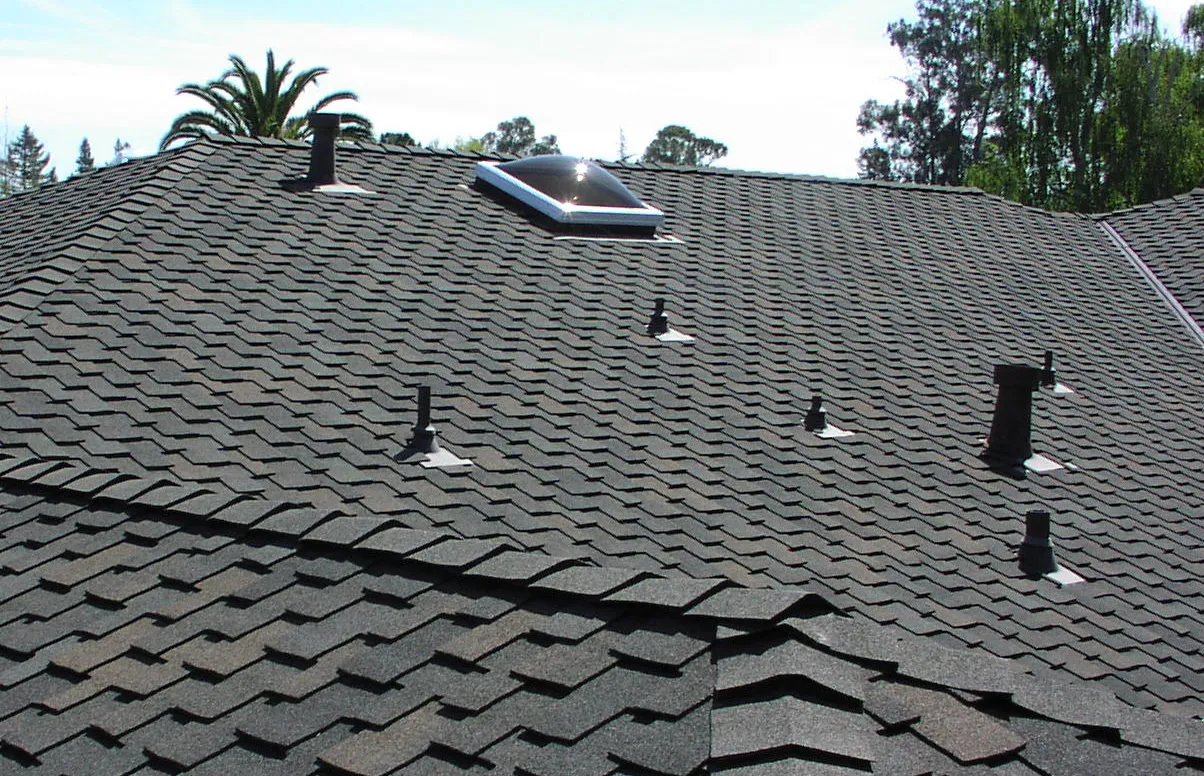 A roof with dark shingles featuring a skylight and several vent pipes. Surrounding trees are visible in the background. A roof with dark shingles featuring a skylight and several vent pipes. Surrounding trees are visible in the background.