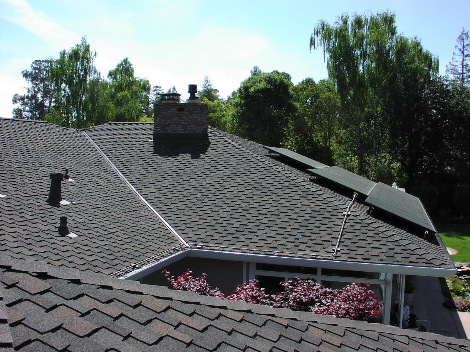 View of a residential roof with dark shingles, a brick chimney, solar panels, and lush trees in the background. View of a residential roof with dark shingles, a brick chimney, solar panels, and lush trees in the background.