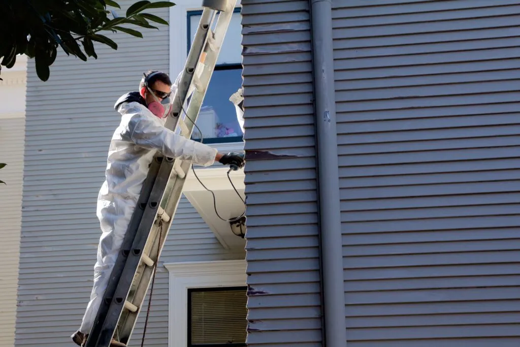 Person wearing protective gear stands on a ladder, working on the siding of a house with a paint scraper. Person wearing protective gear stands on a ladder, working on the siding of a house with a paint scraper.