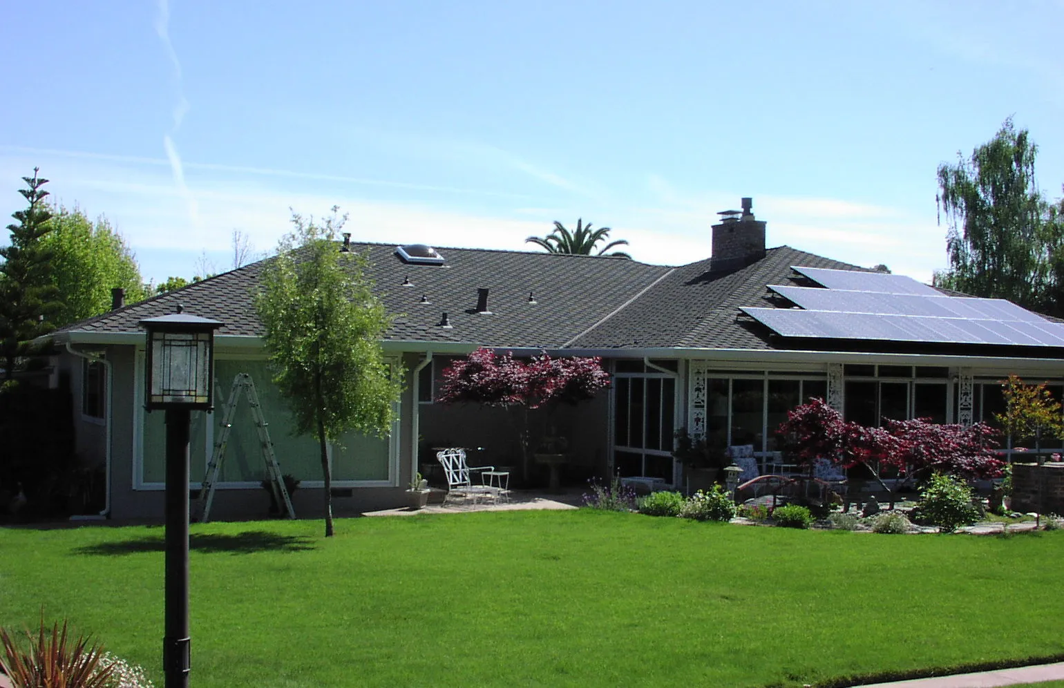 A house with a well-maintained roof features solar panels, a ladder, and a lush garden in the foreground. A house with a well-maintained roof features solar panels, a ladder, and a lush garden in the foreground.