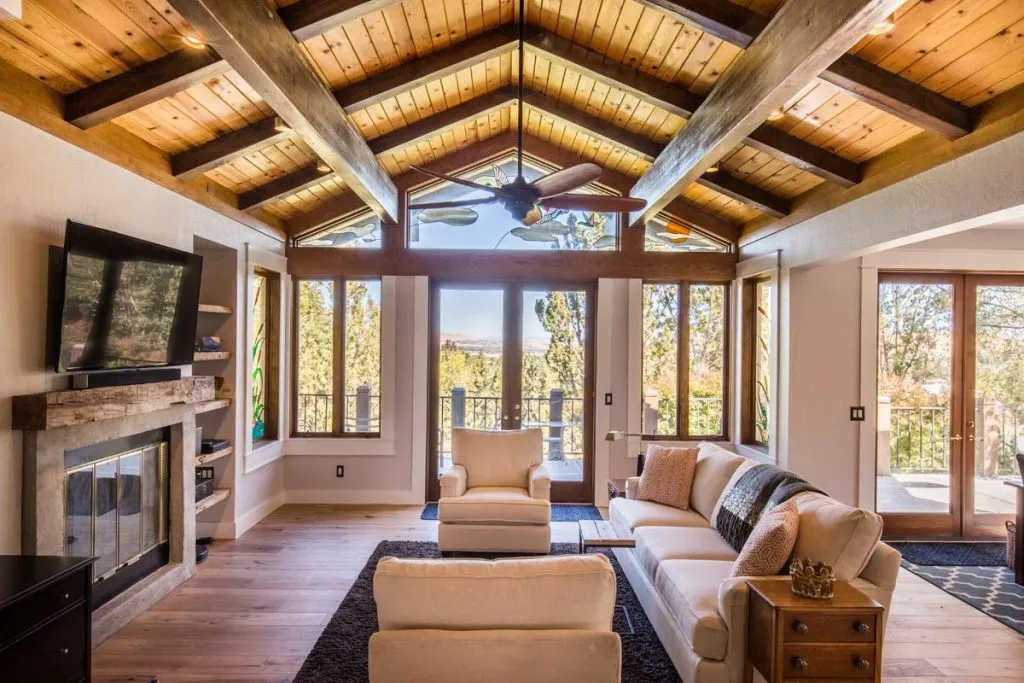 Cozy living room with vaulted wooden ceiling, large windows, white sofas, and a stone fireplace. Natural light fills the space, with trees visible outside. Cozy living room with vaulted wooden ceiling, large windows, white sofas, and a stone fireplace. Natural light fills the space, with trees visible outside.