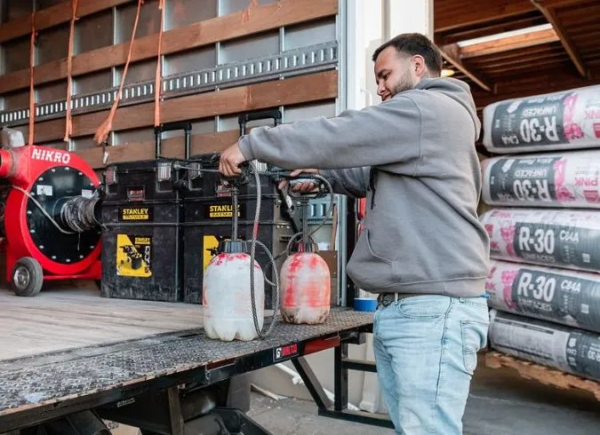 A man in a gray hoodie handles spray equipment on a workshop table, with insulation materials and tools visible in the background. A man in a gray hoodie handles spray equipment on a workshop table, with insulation materials and tools visible in the background.
