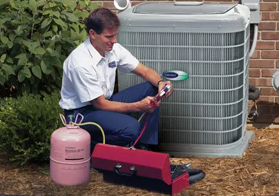  Technician adjusts gauges on an outdoor air conditioning unit with tools and a canister beside him, surrounded by greenery and mulch.