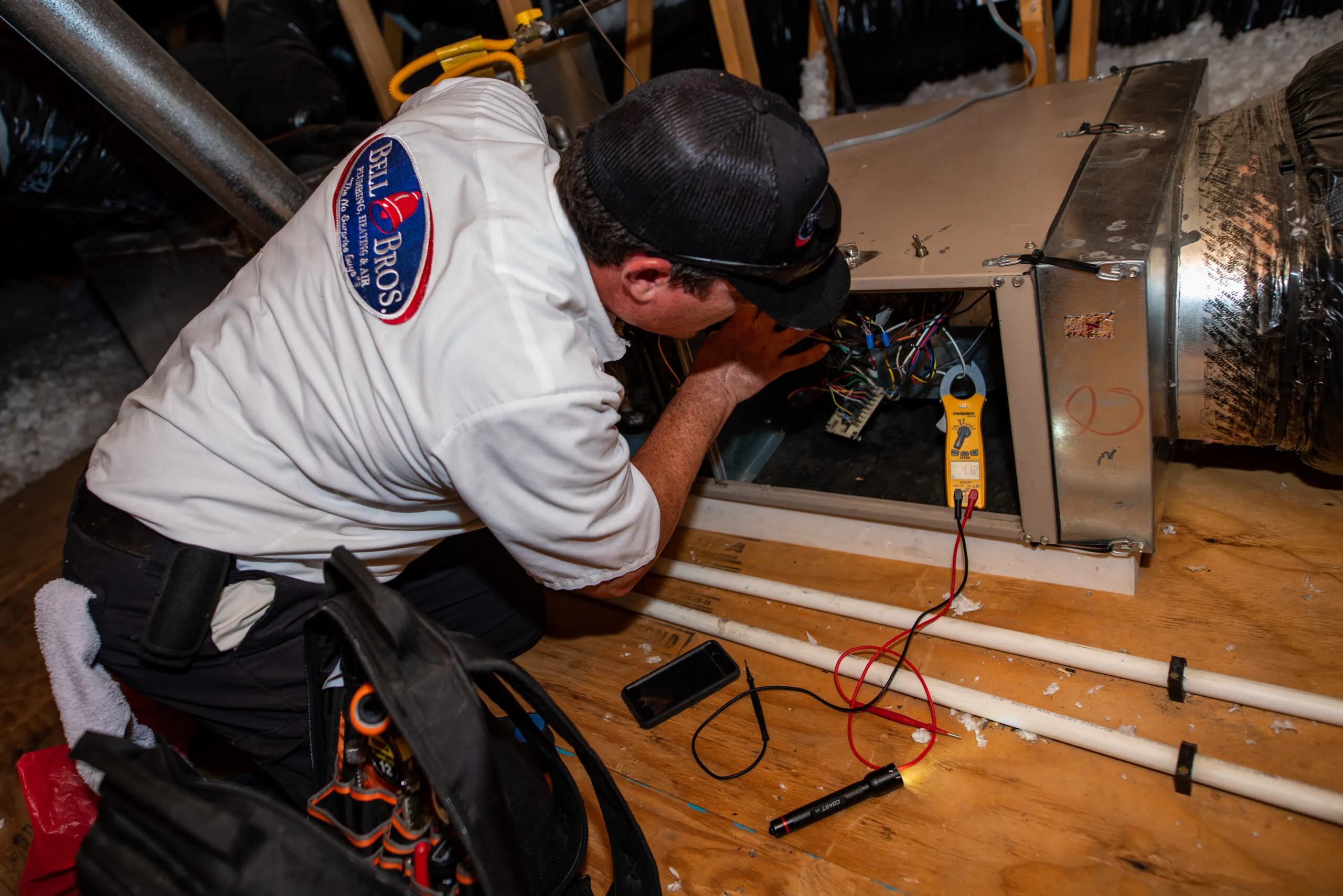  Technician examining HVAC system wiring with tools and equipment on the floor, wearing a Bell Brothers uniform.