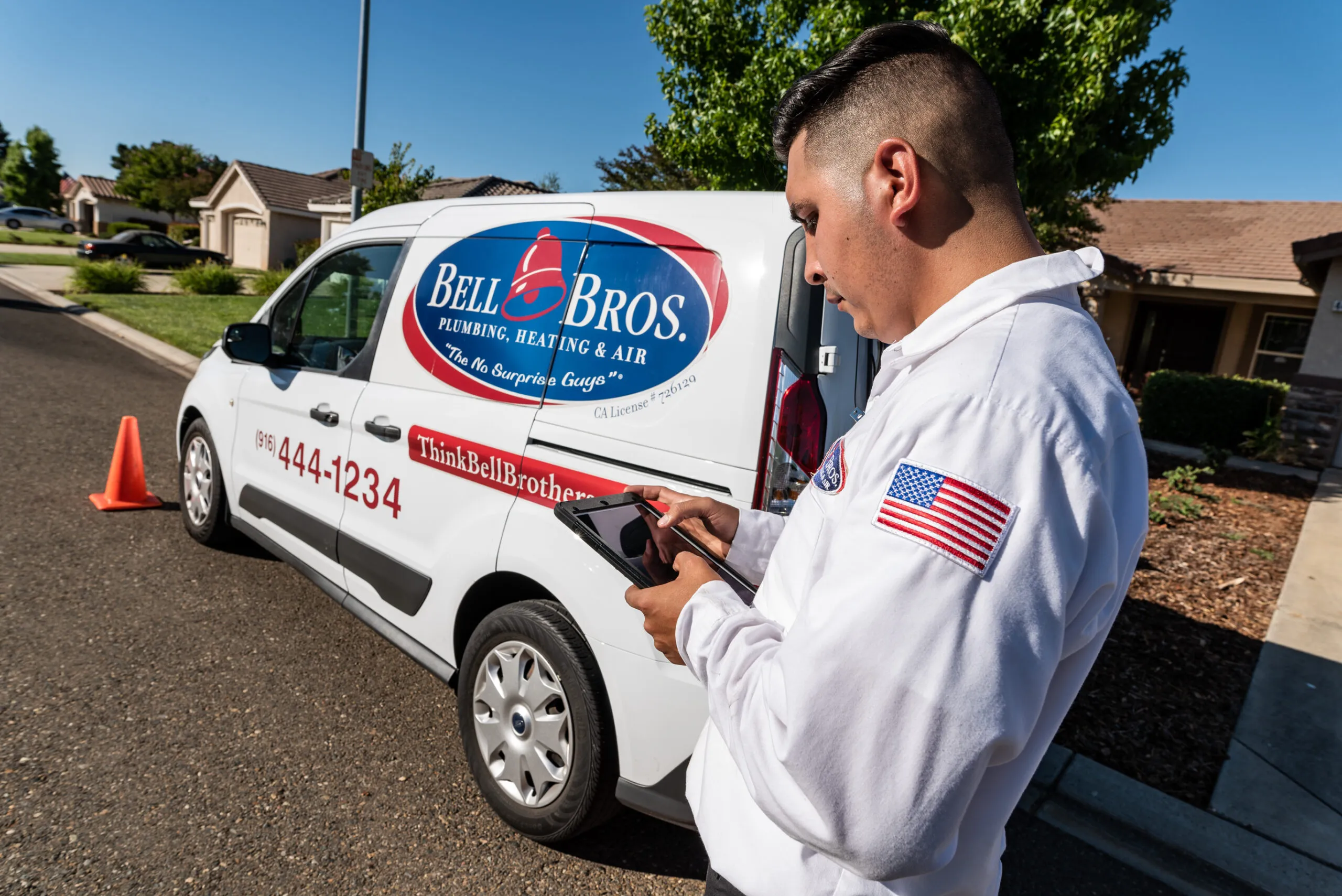  A technician in a white uniform uses a tablet next to a Bell Brothers service van on a residential street. An orange cone is placed near the van.