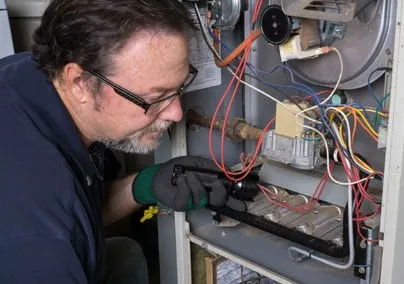  Technician examining the internal components of a furnace, surrounded by wires and mechanical parts, using a flashlight for inspection.