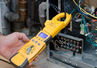A hand holds a yellow clamp meter testing electrical components inside an HVAC unit, surrounded by wires and pipes.