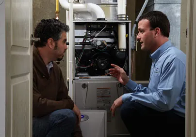 Technician and client kneel beside an open furnace, discussing its components. Technician wears a company-branded shirt, gesturing towards the system.