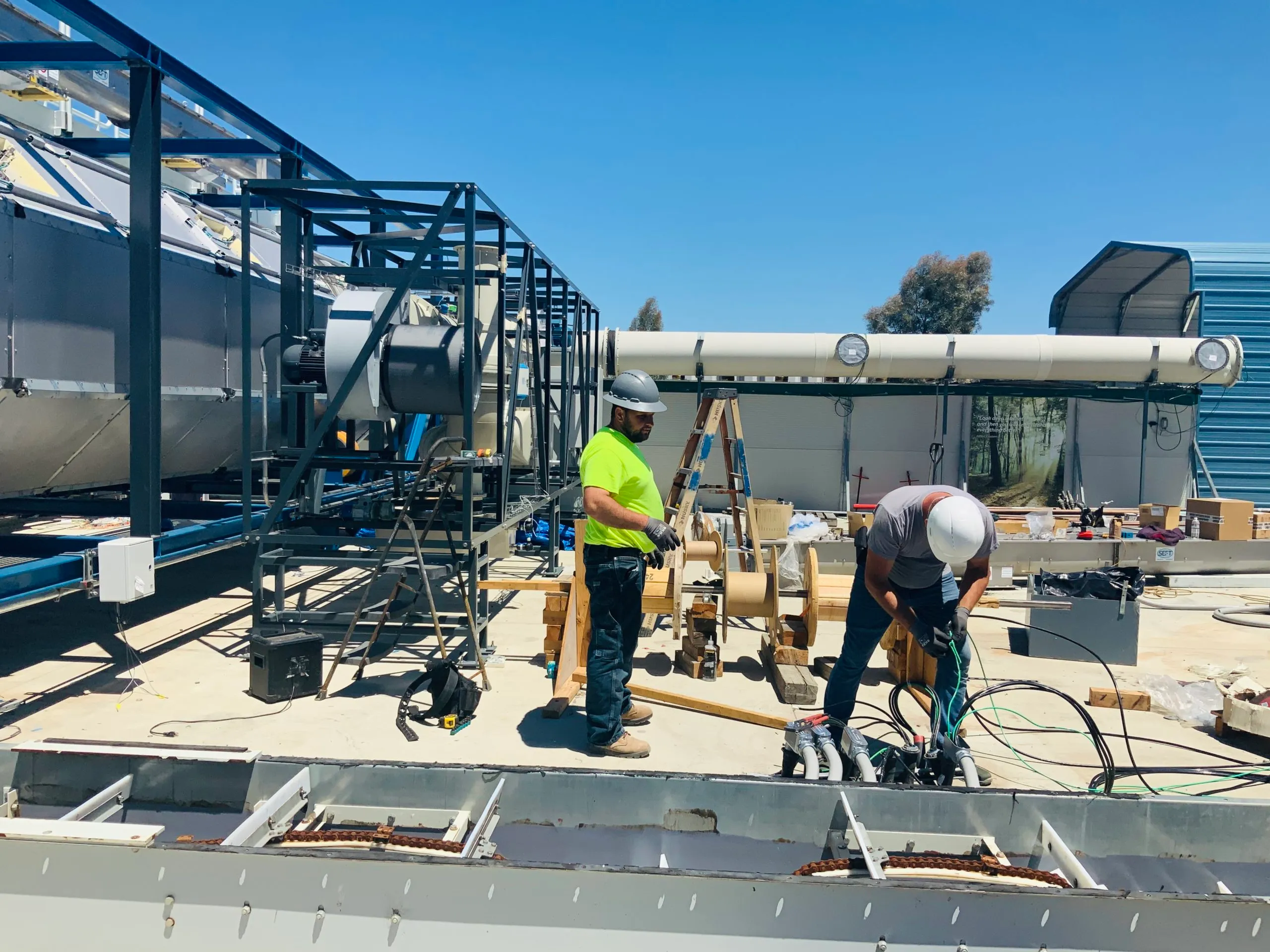  Two workers in safety gear are handling wires on a commercial rooftop. Large industrial equipment and a ladder are nearby, with clear blue skies overhead.