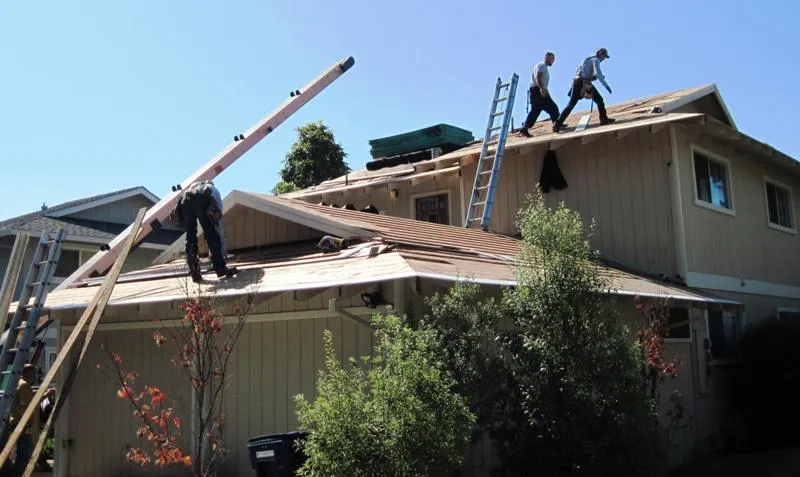 Three workers on ladders and a roof, installing roofing materials on a two-story house with trees in the foreground. Three workers on ladders and a roof, installing roofing materials on a two-story house with trees in the foreground.