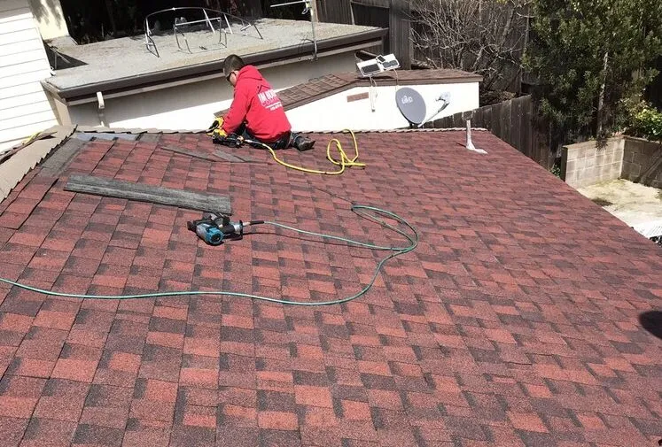 Worker in a red hoodie installs shingles on a red rooftop with tools and hoses nearby. Worker in a red hoodie installs shingles on a red rooftop with tools and hoses nearby.