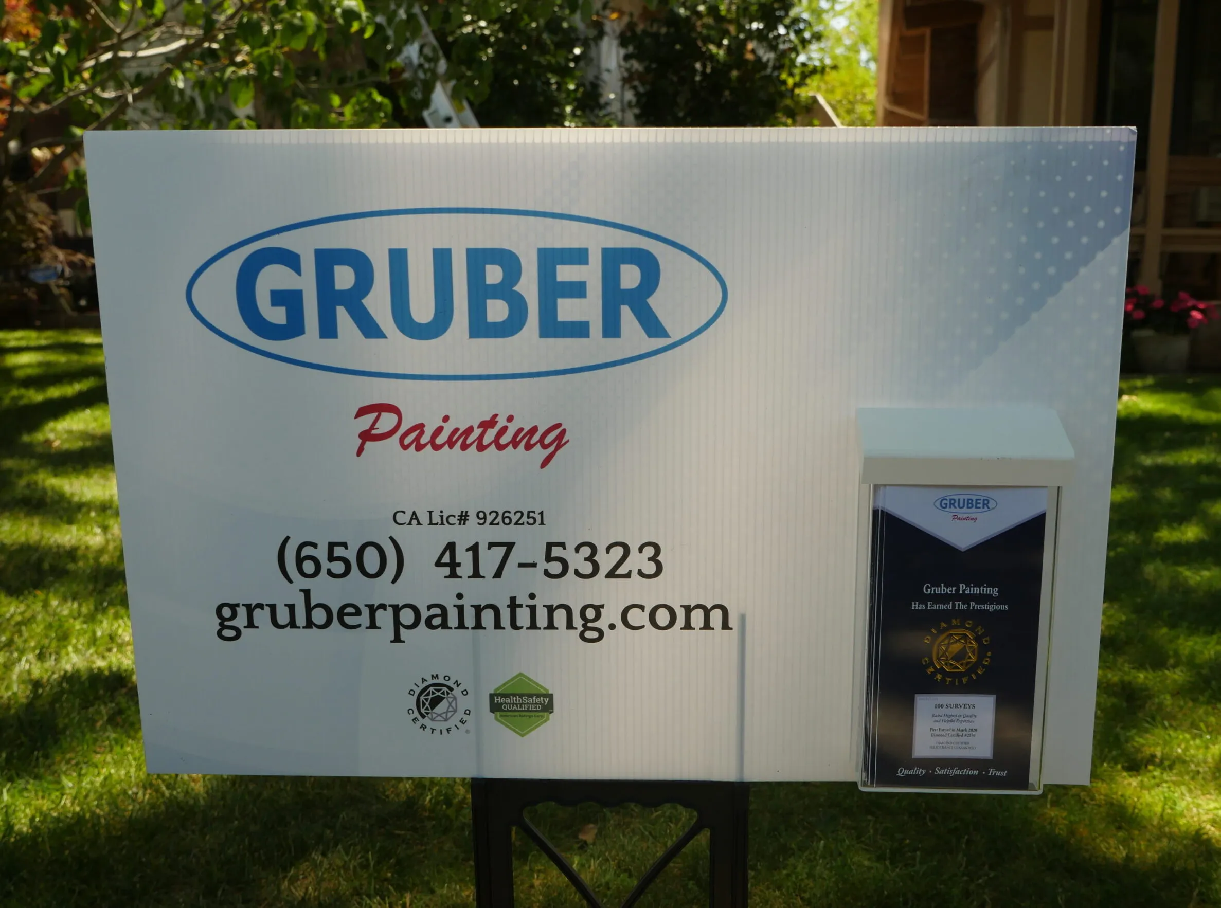 A Gruber Painting sign with contact details and Diamond Certified logo, set in a garden with greenery. A Gruber Painting sign with contact details and Diamond Certified logo, set in a garden with greenery.