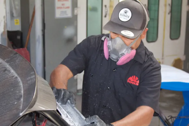 Technician wearing a mask sands a car panel inside a garage, using precision tools. Technician wearing a mask sands a car panel inside a garage, using precision tools.