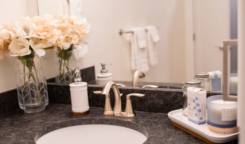 Bathroom sink with floral arrangement, soap dispenser, candles, and toiletries on a dark countertop. White towels hang in the background. Bathroom sink with floral arrangement, soap dispenser, candles, and toiletries on a dark countertop. White towels hang in the background.