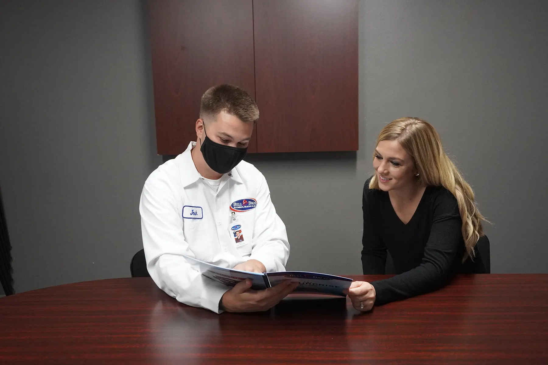 A technician in a Bell Brothers uniform and mask shows a brochure to a woman at a table in an office setting.