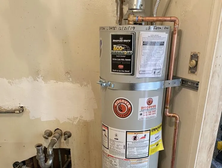  Water heater with Discover Plumbing & Rooter logo, connected copper pipes, and visible installation date in a utility space.